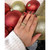 An image of a hand wearing a Rachel Koen unisex adult ring with emeralds, positioned at a slight angle to the camera. The ring is prominently displayed on the ring finger. The background features large, glittery red and gold ornaments, adding a festive touch to the close-up shot.