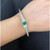 An image of a women's bracelet by Rachel Koen, featuring diamonds and emerald gemstone, positioned on a wrist. The bracelet is angled slightly upward, showcasing its sleek design in white gold. A prominent green gemstone is centrally placed, flanked by diamonds, with the overall view taken from a close distance, highlighting the intricate details. The background is a dark fabric, providing contrast to the bracelet's sparkle.