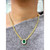 This image shows a gold necklace adorned with a green emerald gemstone in the center, surrounded by round cut diamonds in prong setting. The necklace is being worn over a gray garment.