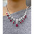 An image of a necklace designed for women, featuring an exquisite arrangement of diamonds and red rubies. The necklace is worn around the neck and displayed against a grey fabric background. The diamonds form floral patterns, with red teardrop rubies hanging elegantly along the piece in white gold. The photograph is taken from a close-up, frontal angle, highlighting the intricate details and sparkling elements of the jewelry.
