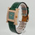 An image of a pre-owned Hermès women's wristwatch displayed at a slight angle, positioned to the right. The watch features a green leather strap and a square face with a green dial. The rose gold case is adorned with sparkling stones, and the watch is mounted on a transparent stand, showcasing its elegant design from a close-up perspective.