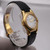 An image of a pre-owned Patek Philippe women's wristwatch, showcasing a yellow gold case and a black leather strap. The watch face is angled slightly to the right, displaying Roman numeral hour markers and a date indicator. The watch is positioned in the center foreground, with the strap extending towards the background. The photo is taken from a close-up perspective, highlighting the intricate details and elegant design.