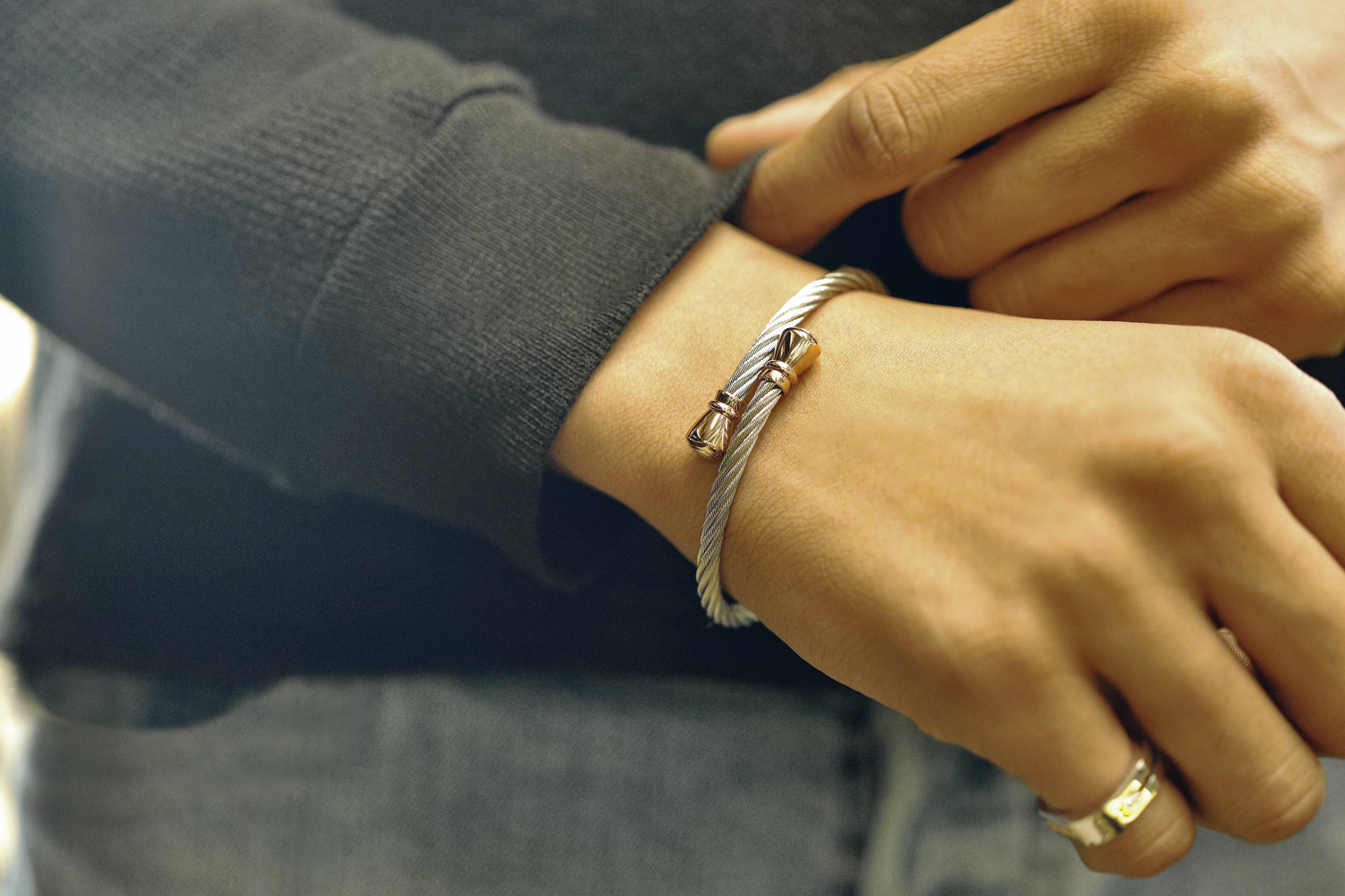 Image of a man wearing blue jeans and a black long sleeve shirt holding his sleeve's cuff to  adjust it around a Charriol bangle that resembles wound steel cables as that of a bridge or rope with rose gold ends.