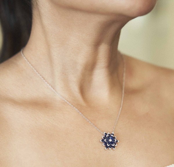 An image of a woman with volumous brown hair wearing a blazer with a diamond and pearl encrusted yellow gold necklace and match yellow gold earrings. 