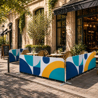 A colourful sidewalk cafe barrier featuring blue, yellow, and white geometric patterns, with tables and chairs visible behind it.