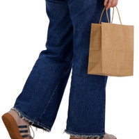 A medium brown paper bag held by a person wearing blue jeans and casual shoes. The bag has handles for easy carrying.