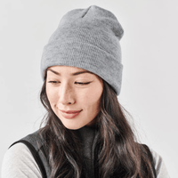 A grey knit beanie worn by a woman with long, wavy hair, paired with a dark vest. It features a logo.