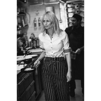 A woman in a white button-up shirt with black trim walks through a kitchen holding a frying pan, wearing striped pants.
