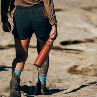 Woman walking on rocky terrain, wearing shorts and hiking boots, holding a red Camelbak water bottle, showcasing colorful leg tattoos.