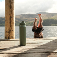 Person jumping of a wooden jetty into a lake while a green Camelbak stainless steel water bottle is stading on the jetty waiting for the person.