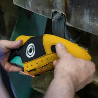 A close-up of hands holding a climbing shoe as it undergoes finishing on a grinding wheel in a workshop.