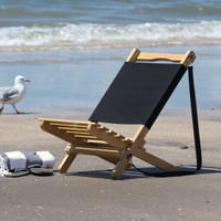 A wooden beach chair with a black fabric seat sits on the sand near the water. Two rolled towels are beside it.