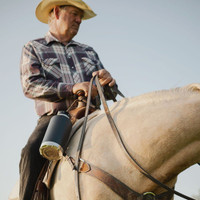 An elderly gentleman is sitting on an palomino horse. The horse is only partially visible. A black Fridgy 1.9l All-Dayer is tied to the saddle.