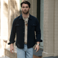 A unisex shirt in navy with buttons, worn over a beige top, paired with light jeans. The model is walking outdoors.