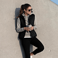 A woman in a black vest over a grey hoodie, holding a drink, leaning against a concrete wall, wearing black leggings and white shoes.