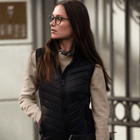A woman in a black vest over a camel brown jumper, her right hand in the pocket of the vest and hodling a black document pouch in the left hand.