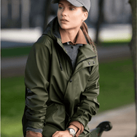 A green unisex jacket is worn by a woman sitting on a bench, paired with a grey cap and neutral trousers.