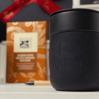A black branded mug with a logo in the foreground, set against a patterned gift box and a pen.