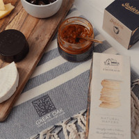 A wooden board with cheese, a jar of spread, and a box of wafers on a textured cloth with a logo.