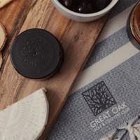 A wooden platter featuring a round black cheese, olives, and a slice of cheese, alongside a textured cloth with branding.