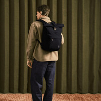 A person wearing a black Bellroy Classic Rolltop backpack, standing against a green curtain backdrop.