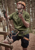 A man wearing a green short sleeve button-up shirt and blue shorts, equipped with climbing gear in a forest setting.