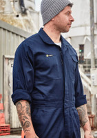 A man wearing a navy blue lightweight cotton drill overall with a logo on the chest, standing in an industrial setting.