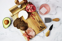 A wooden cheeseboard with cheese, meats, avocado, bread, and a pomegranate, featuring utensils and a logo.