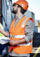 A man wearing an orange hi-vis zip vest with reflective stripes and a hard hat, holding documents at a construction site.