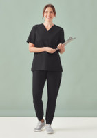 A woman wearing a black V-neck scrub top and black pants, holding a clipboard against a light green background.