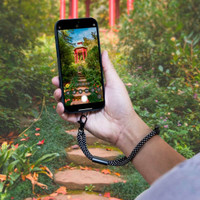 A smartphone is held by a hand with a black and white polka dot wrist strap, set against a lush garden background.