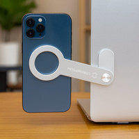 A silver magnetic phone holder attaches a blue smartphone to a laptop. The product has a brand logo.