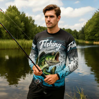 A man wearing a long-sleeved, blue and black sublimated fishing shirt stands by a river holding a fishing rod.