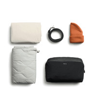 A cream blanket, grey quilted item, black packing cube, an orange beanie, and a black belt, all neatly arranged.