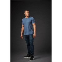 A crew neck t-shirt in blue, displayed on a male model against a dark background, paired with dark jeans and sneakers.