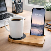 A white mug sits on a bamboo cup warmer with a phone charging beside it. The setting includes a laptop and office supplies.