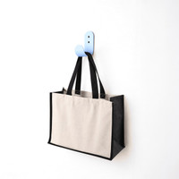 A functional tote bag in beige and black hangs on a blue hook against a white wall. The bag has handles and a logo.