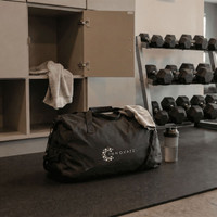 A black duffel bag with a logo, resting on a table alongside a drink bottle and a towel, in a gym setting.