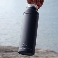 A drink bottle in matte black with a silver cap, featuring a logo and held over a body of water.