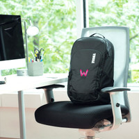A black Thule Achiever 15 laptop backpack with a logo resting on a chair beside a desk and computer.