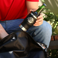 A drink bottle tumbler in black with a silver base, holding a green bottle, placed beside a black bag.