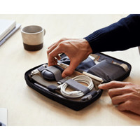 A black tech kit with compartments for cables, a mouse, and accessories, being held by a hand on a table.