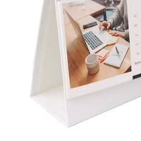 A desk calendar in a white stand, featuring a photo of a workspace with a laptop, notebook, and cup.