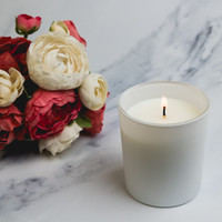 A large white glass candle with a lit wick beside a bouquet of cream and pink flowers.