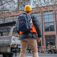 A person wearing a yellow hard hat and an orange vest carries a navy blue High Sierra Access 3.0 Backpack.