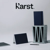 A navy hardcover notebook stands on top of a grey block, accompanied by a smaller notebook and a pen. It has a logo.