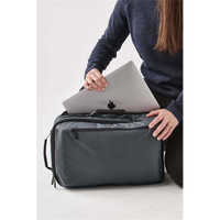 A grey laptop backpack with a person placing a silver laptop inside, featuring a front pocket and a logo.