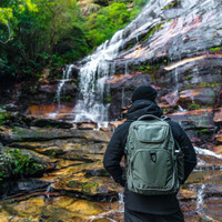 A person stands by a waterfall, wearing a green High Sierra Access 4.0 backpack. The surroundings are lush and rocky.