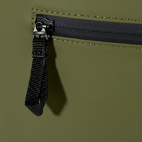 A close-up of a dark olive green roll-top backpack featuring a zippered pocket and a black zipper pull.