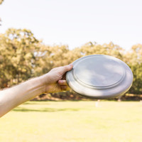 A grey frisbee is held in a hand against a blurred outdoor background. It has a simple design and a logo.